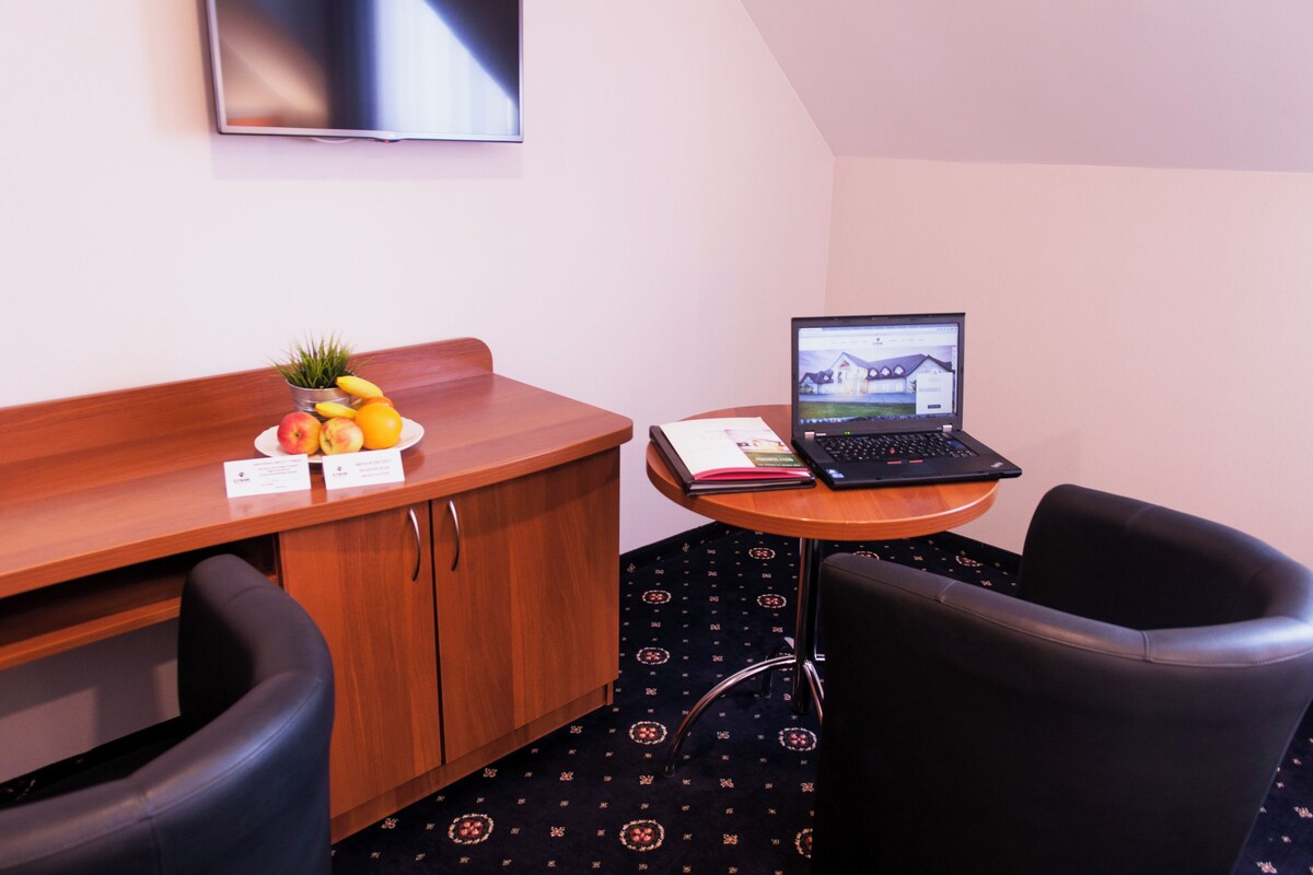 A hotel room with a wooden desk, a laptop, a small table with a chair, a fruit bowl, and a television mounted on the wall.