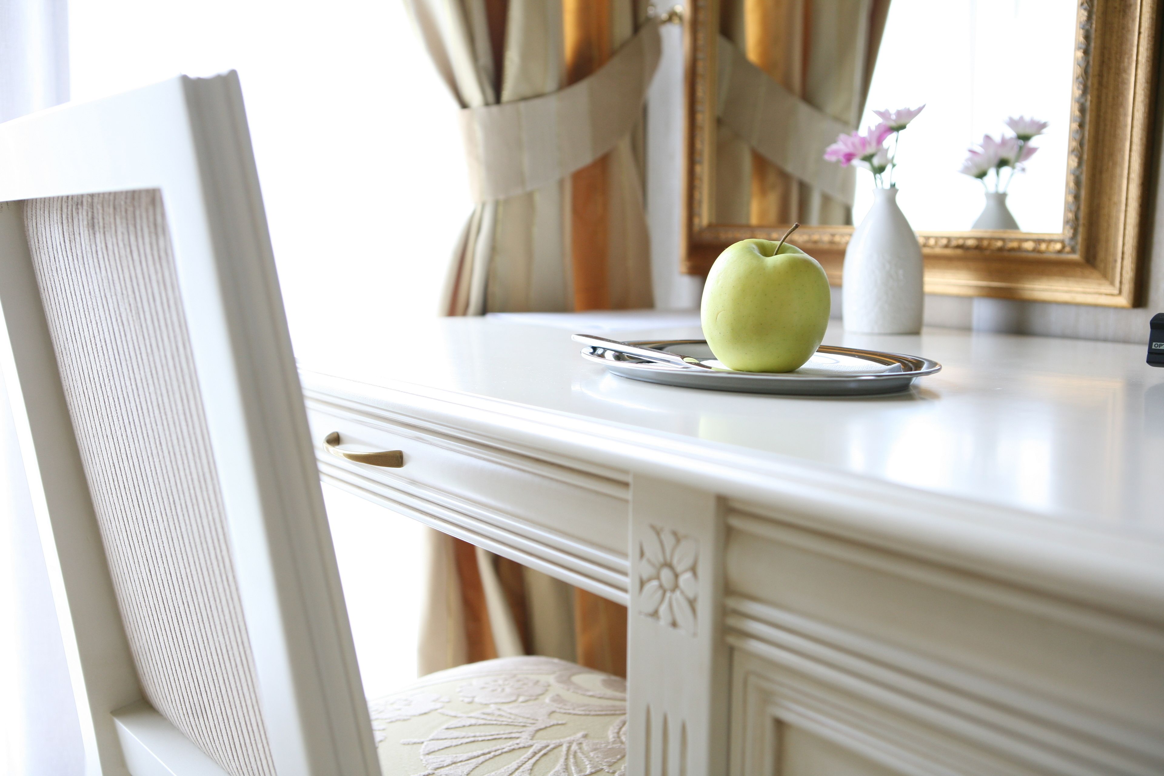 A white table with a green apple on a tray, a white vase with flowers, and a mirror with a gold frame.