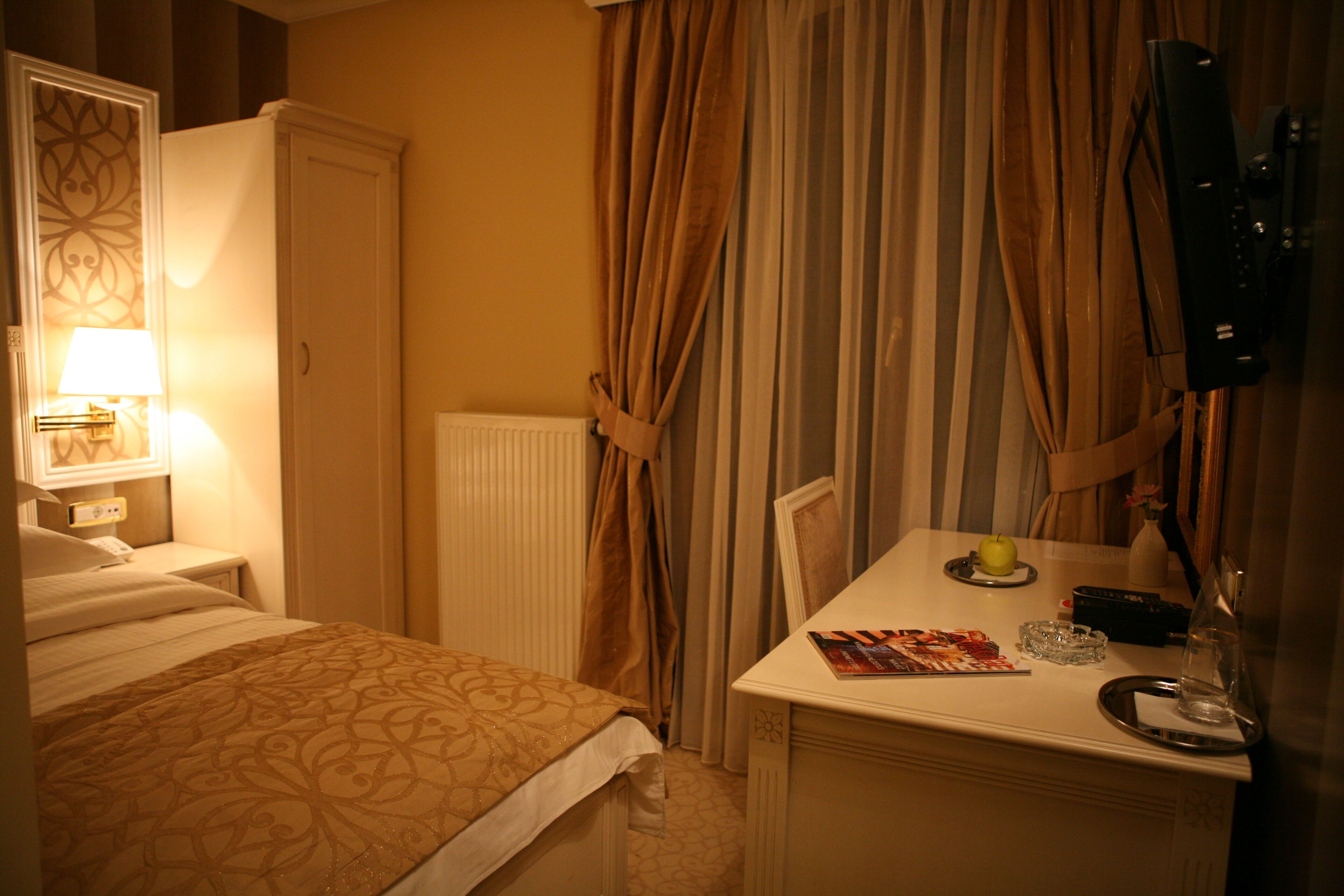 Une chambre d’hôtel avec un lit, une table de chevet, une lampe, des rideaux et une petite table sur laquelle se trouvent un magazine, un verre et une bouteille.