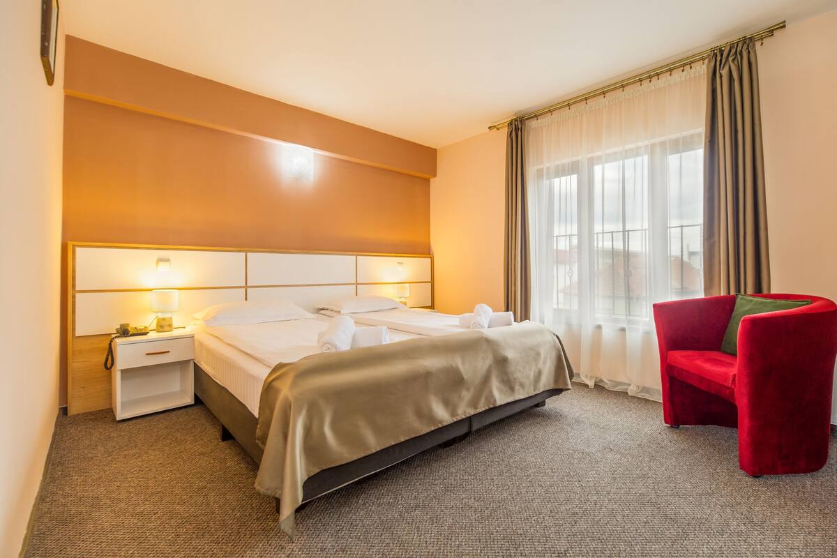 A hotel room with a bed, a nightstand, a red armchair, and a window with curtains.