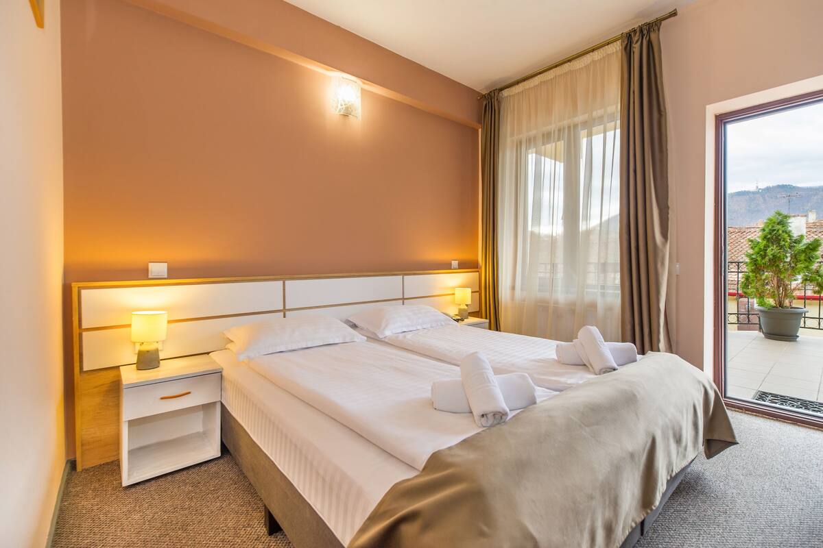 A hotel room with a large bed, two bedside tables with lamps, a view of the city through a balcony door.