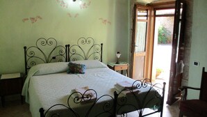 A bedroom with a metal bed frame, a bedside table, a nightstand, a window with curtains, and a door leading outside. - Sa Dom'e Forru (Collinas)