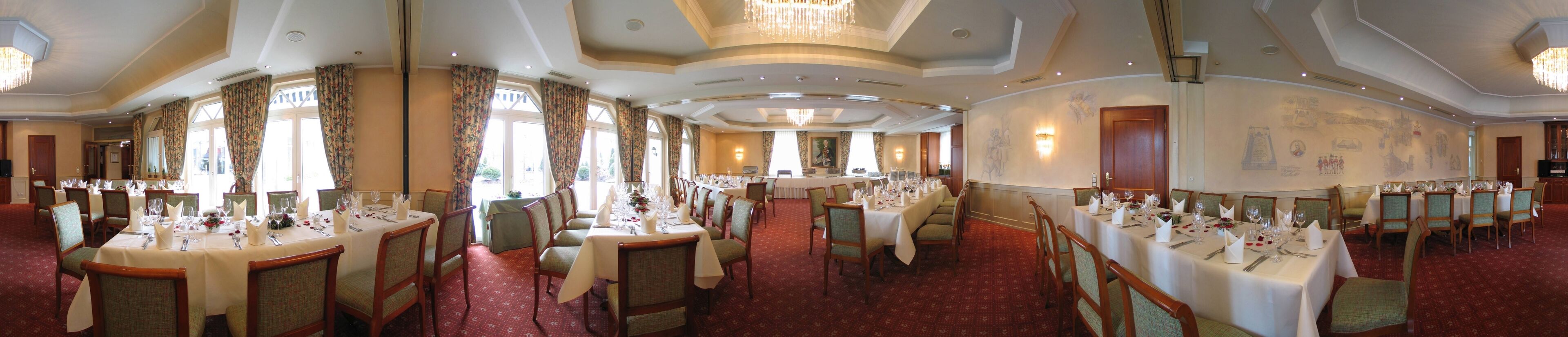 A large banquet hall with multiple round tables set for dining, high ceilings with chandeliers, and large windows with curtains.