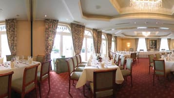 A large banquet hall with multiple round tables set for dining, high ceilings with chandeliers, and large windows with curtains.