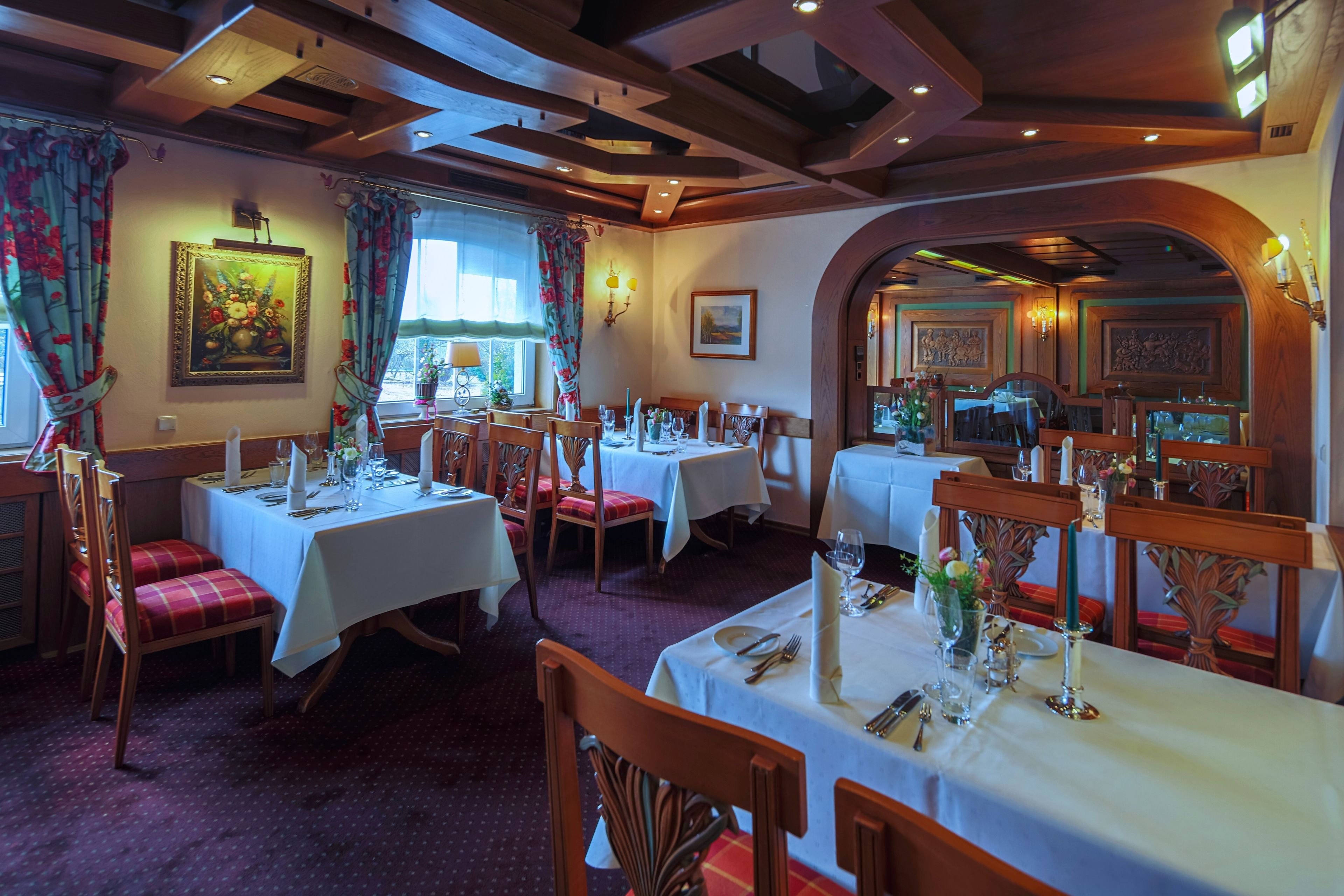 A restaurant with multiple tables set for dining, wooden chairs, and a warm interior with wooden beams on the ceiling.