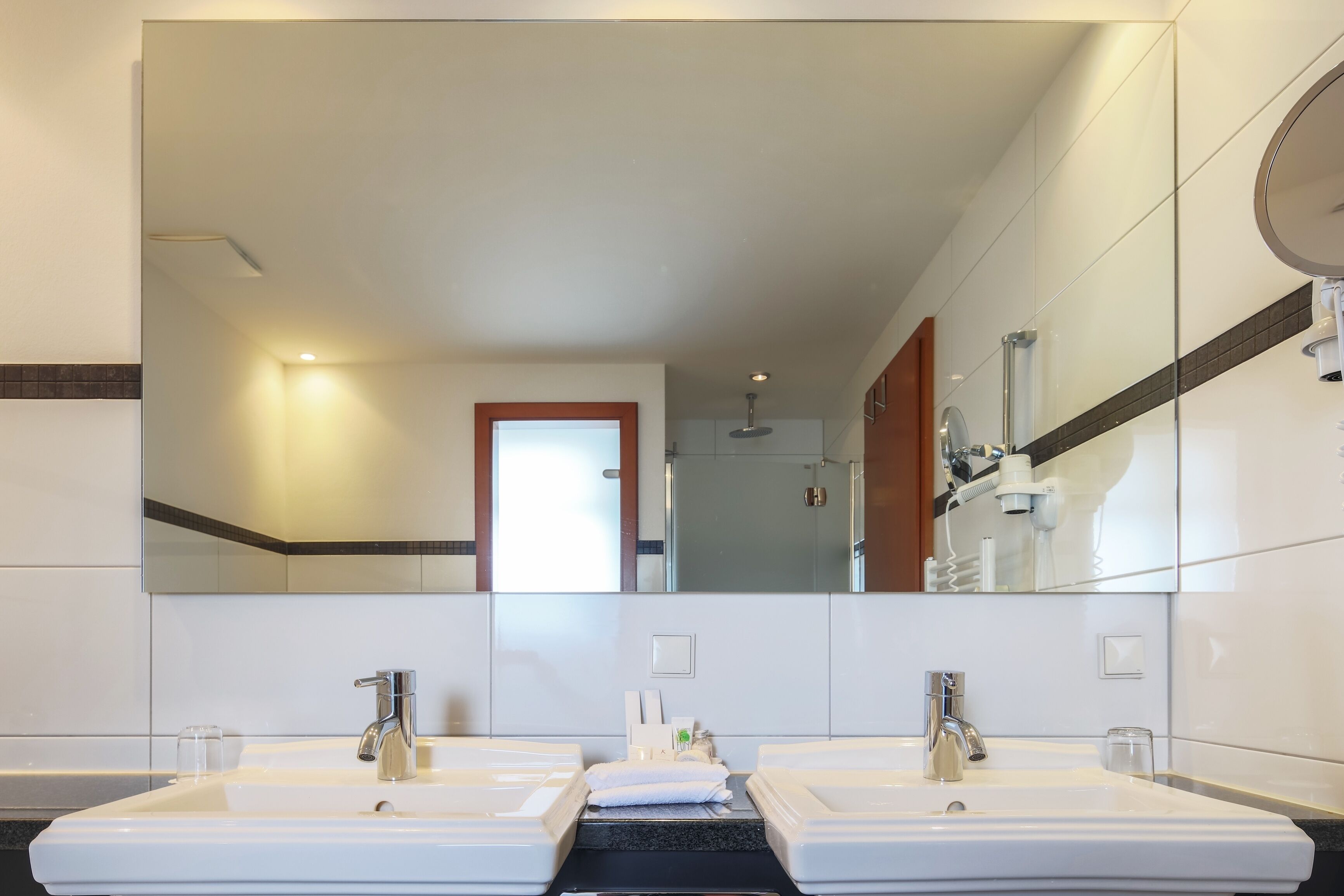 A hotel bathroom with two sinks, a large mirror, and a shower visible in the background.