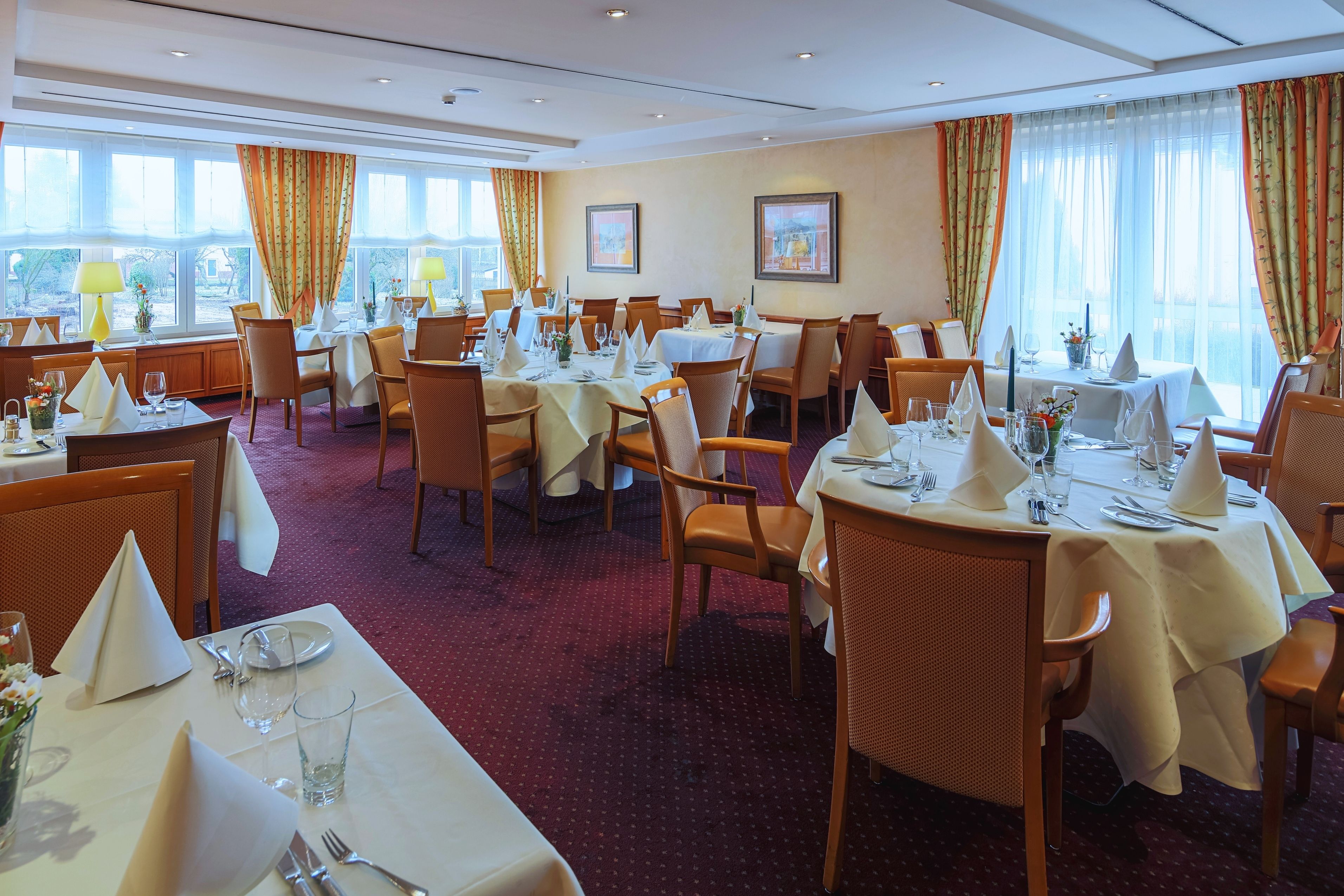 Un comedor de hotel con varias mesas dispuestas para los huéspedes, ventanas amplias con cortinas y obras de arte enmarcadas en las paredes.