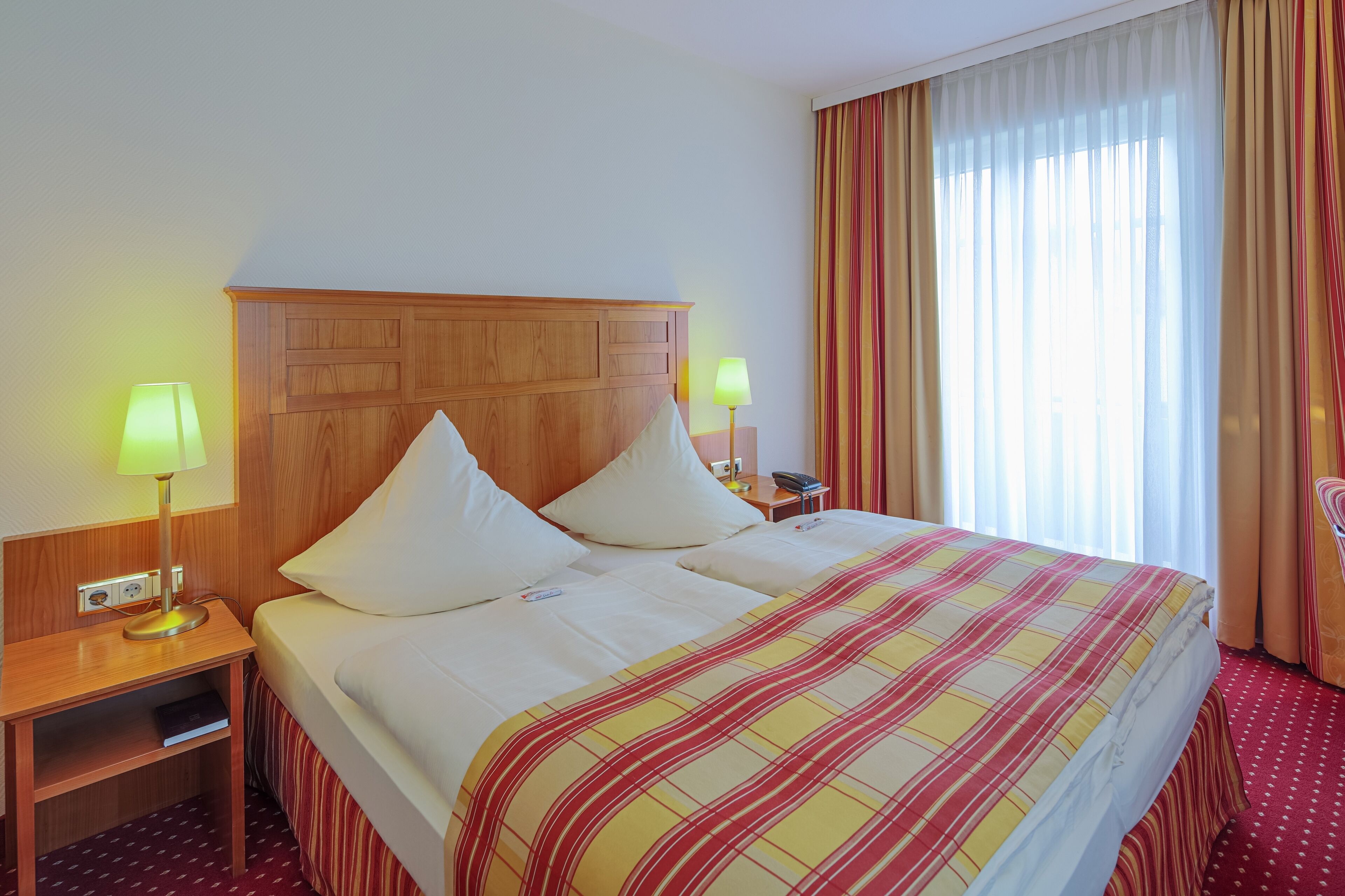 A hotel room with a large bed, two bedside lamps, a wooden headboard, and a window with curtains.