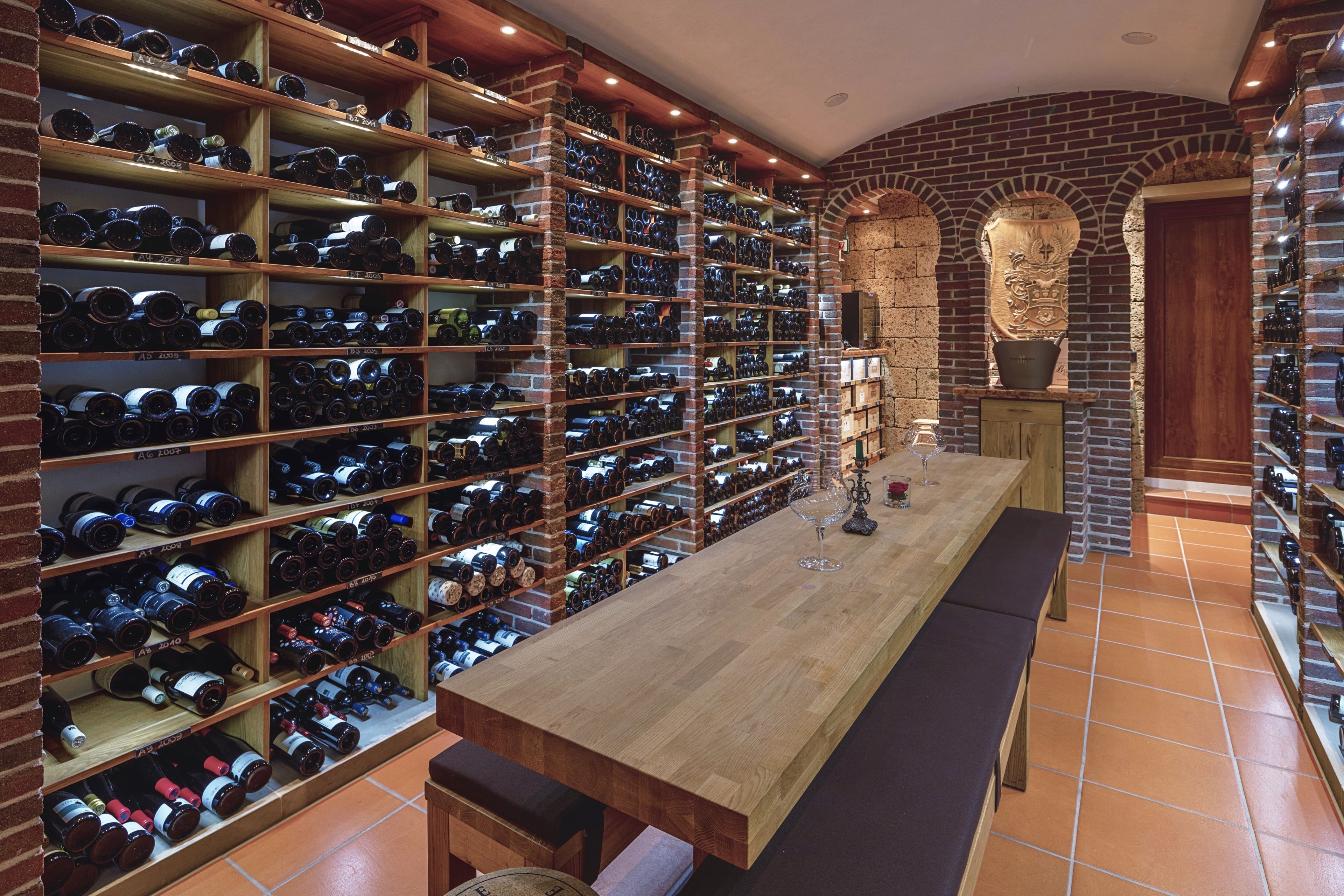 A wine cellar with wooden shelves and a long table, featuring a brick wall and arched doorways.