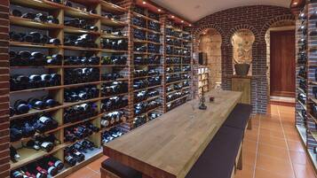 A wine cellar with wooden shelves and a long table, featuring a brick wall and arched doorways.