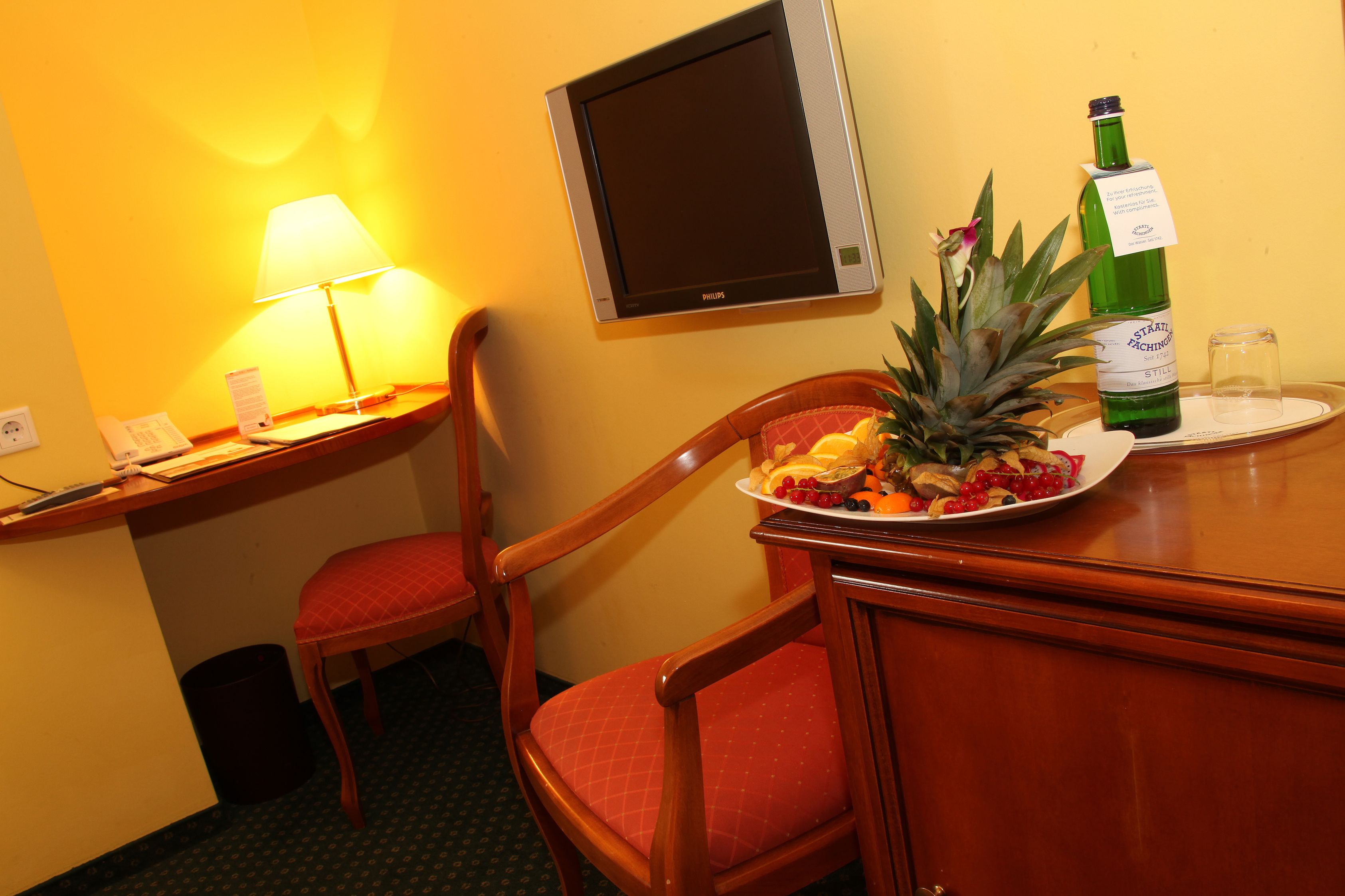Een hotelkamer met een houten bureau, een stoel, een televisie, een fles wijn en een schaal fruit.