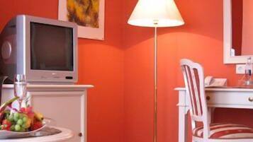 Une chambre équipée d’une télévision, d’un bureau, d’une chaise et d’une table sur laquelle se trouvent une bouteille de champagne et des fruits.