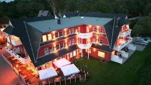 Un edificio grande de varios pisos con fachada roja y techo oscuro. Cuenta con varios balcones y un espacio exterior bien iluminado con toldos blancos.