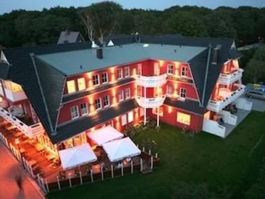 A large, multi-story building with a red facade and a dark roof. It features multiple balconies and a well-lit outdoor area with white canopies.