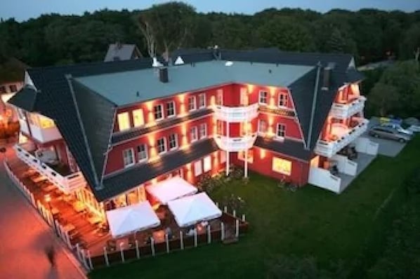 Un edificio grande de varios pisos con fachada roja y techo oscuro. Cuenta con varios balcones y un espacio exterior bien iluminado con toldos blancos.