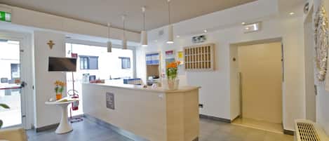 A reception area with a light-colored reception desk, a small white table with a plant, and a chair. There are ceiling lights, a wall-mounted TV, and a door leading outside.
