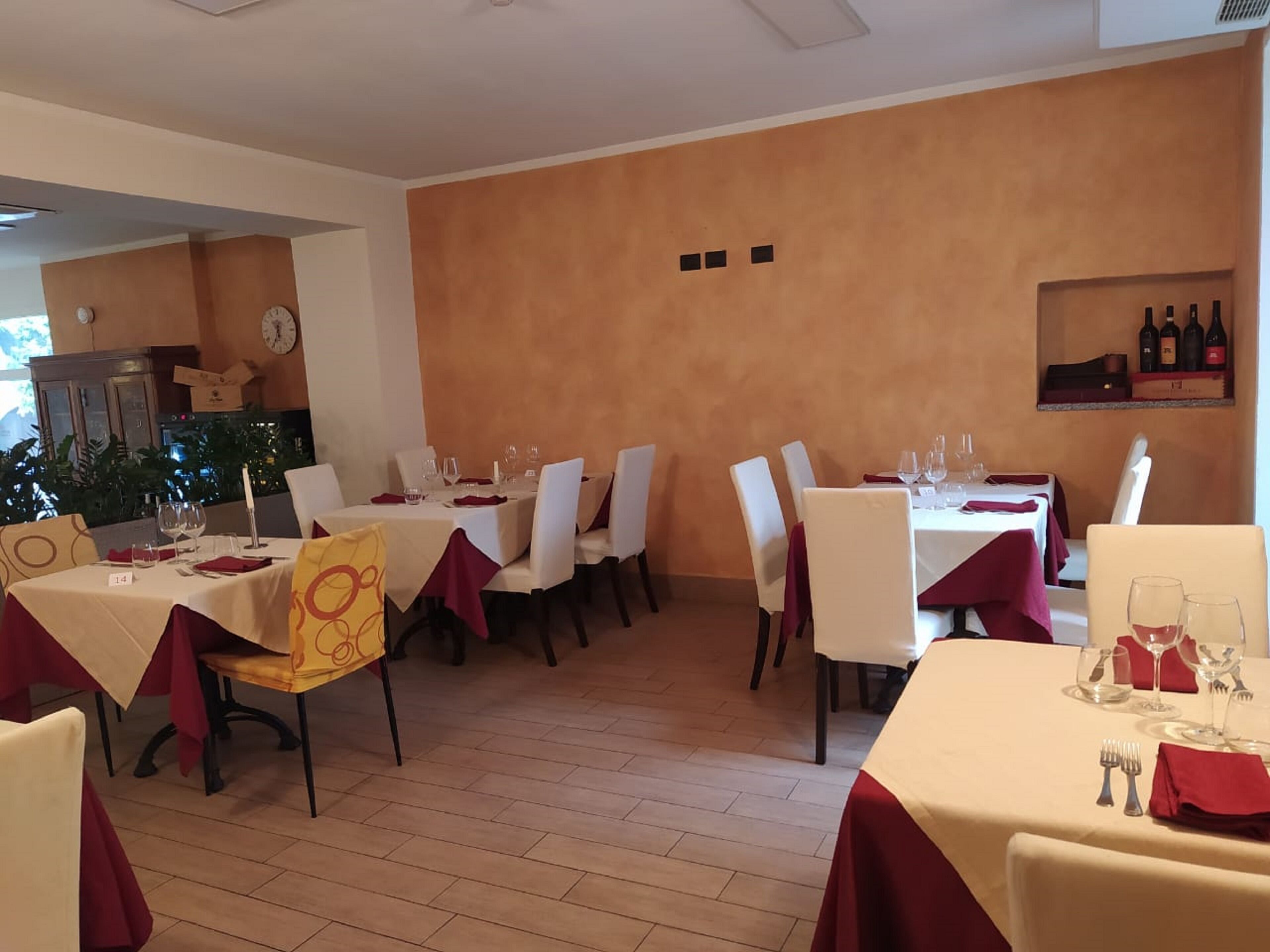 Un restaurante con varias mesas, cada una cubierta con manteles blancos y servilletas rojas. Las paredes están pintadas en un tono naranja cálido, y hay botellas de vino exhibidas en una estantería.