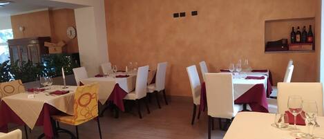 Un restaurante con varias mesas, cada una cubierta con manteles blancos y servilletas rojas. Las paredes están pintadas en un tono naranja cálido, y hay botellas de vino exhibidas en una estantería.