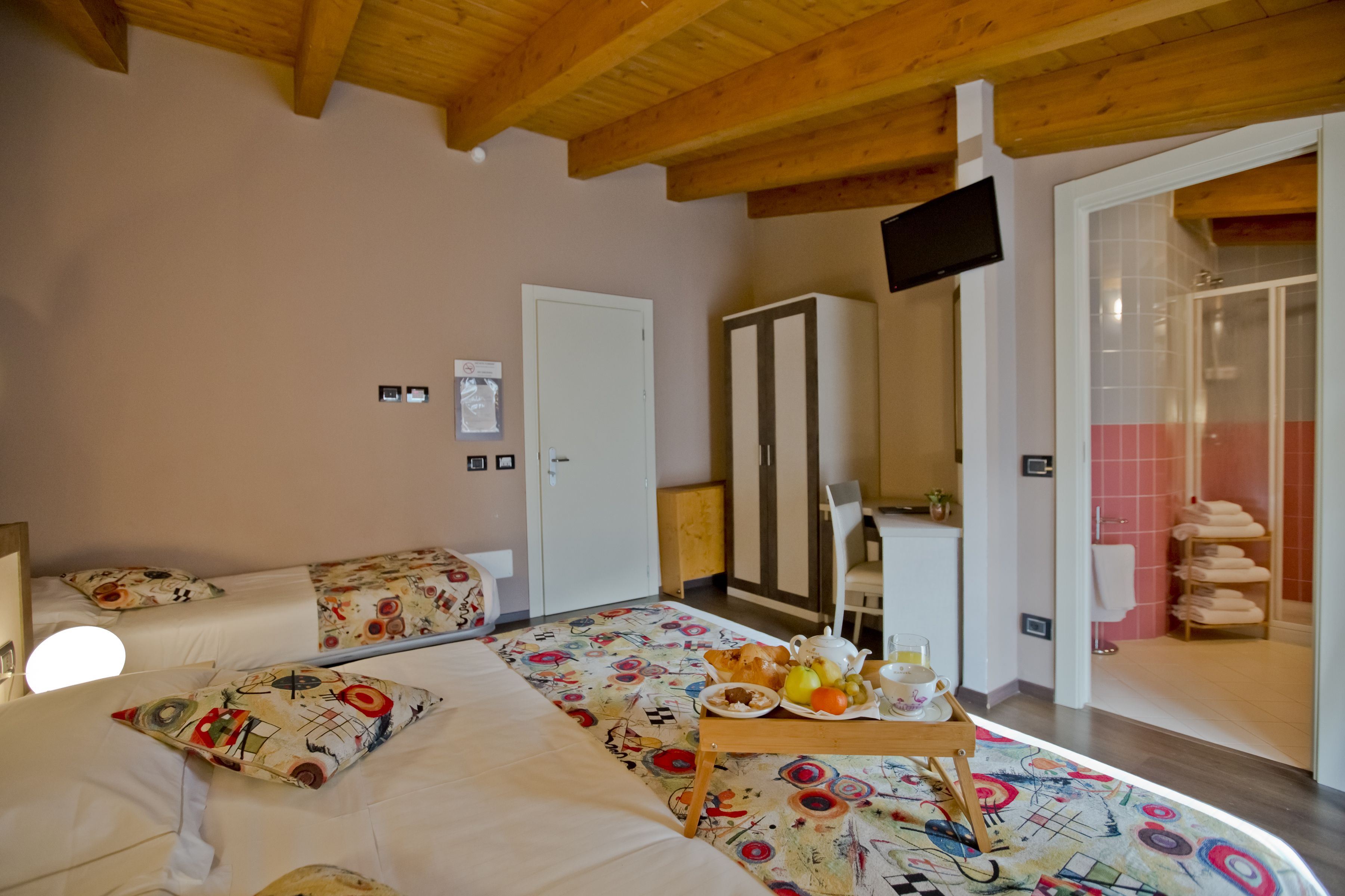 Ein Hotelzimmer mit Holzboden, einem Bett mit gemusterten Betttextilien, einem kleinen Tisch mit Frühstück, einem an der Wand montierten Fernseher und einem durch eine geöffnete Tür einsehbaren Badezimmer.
