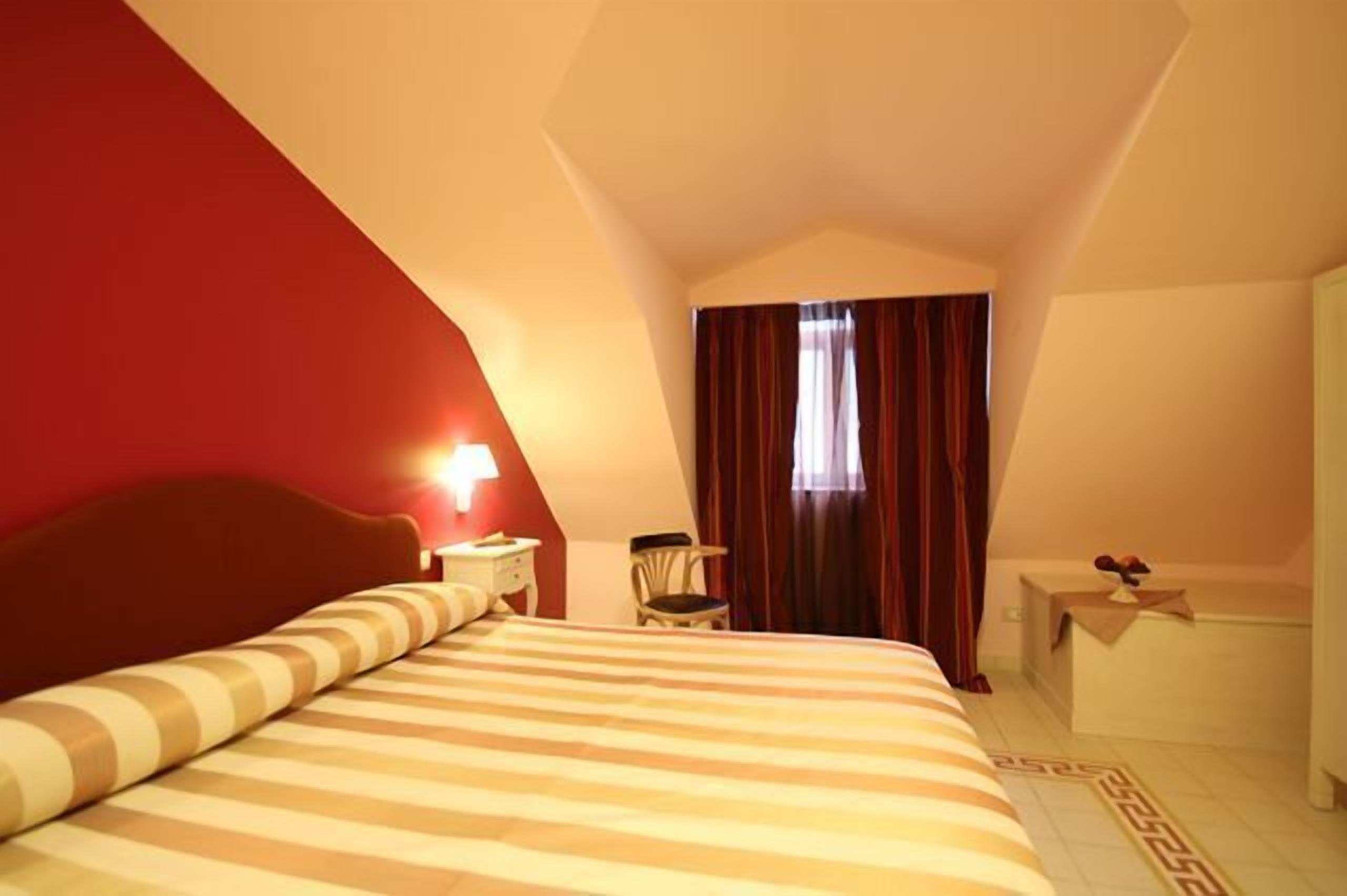 Una habitación de hotel con una cama grande, una mesita de noche, una silla, una ventana con cortinas y una pequeña mesa con lámpara.