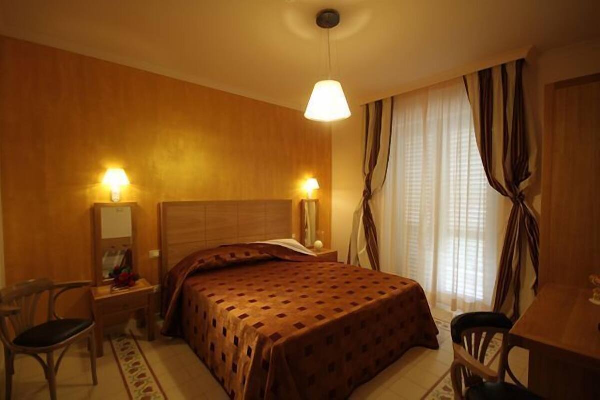 A hotel room with a bed, a desk, a chair, a lamp, and curtains.