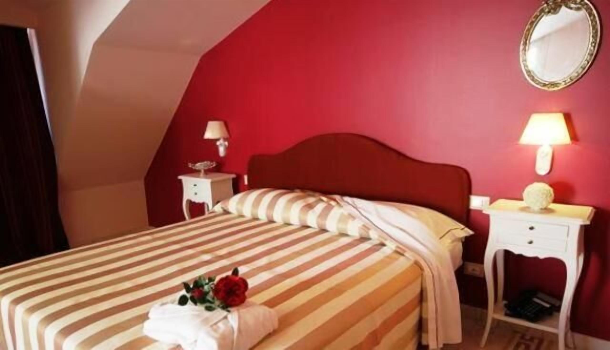 Una habitación de hotel con una pared de acento roja, una cama con ropa de cama a rayas, dos mesitas de noche con lámparas y un espejo en la pared.