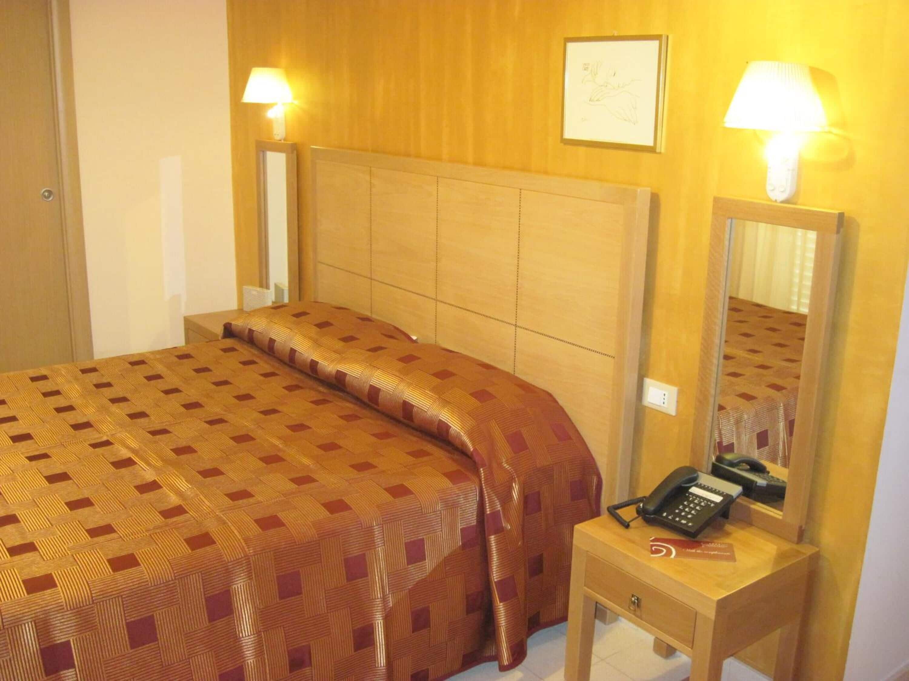 A hotel room with a bed, a bedside table with a telephone, and wall-mounted lamps.