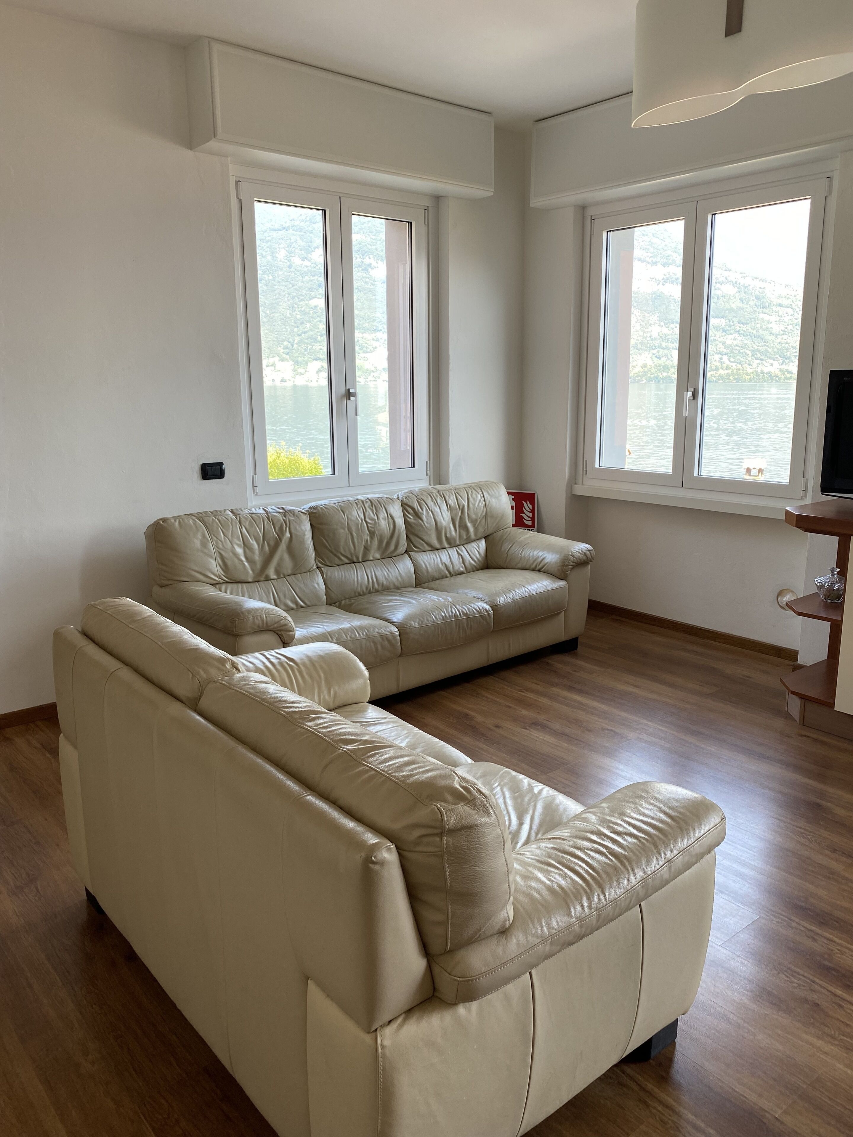 En rummelig stue med en stor, L-formet sofa i beige læder, to vinduer med udsigt til en sø, og trægulv.