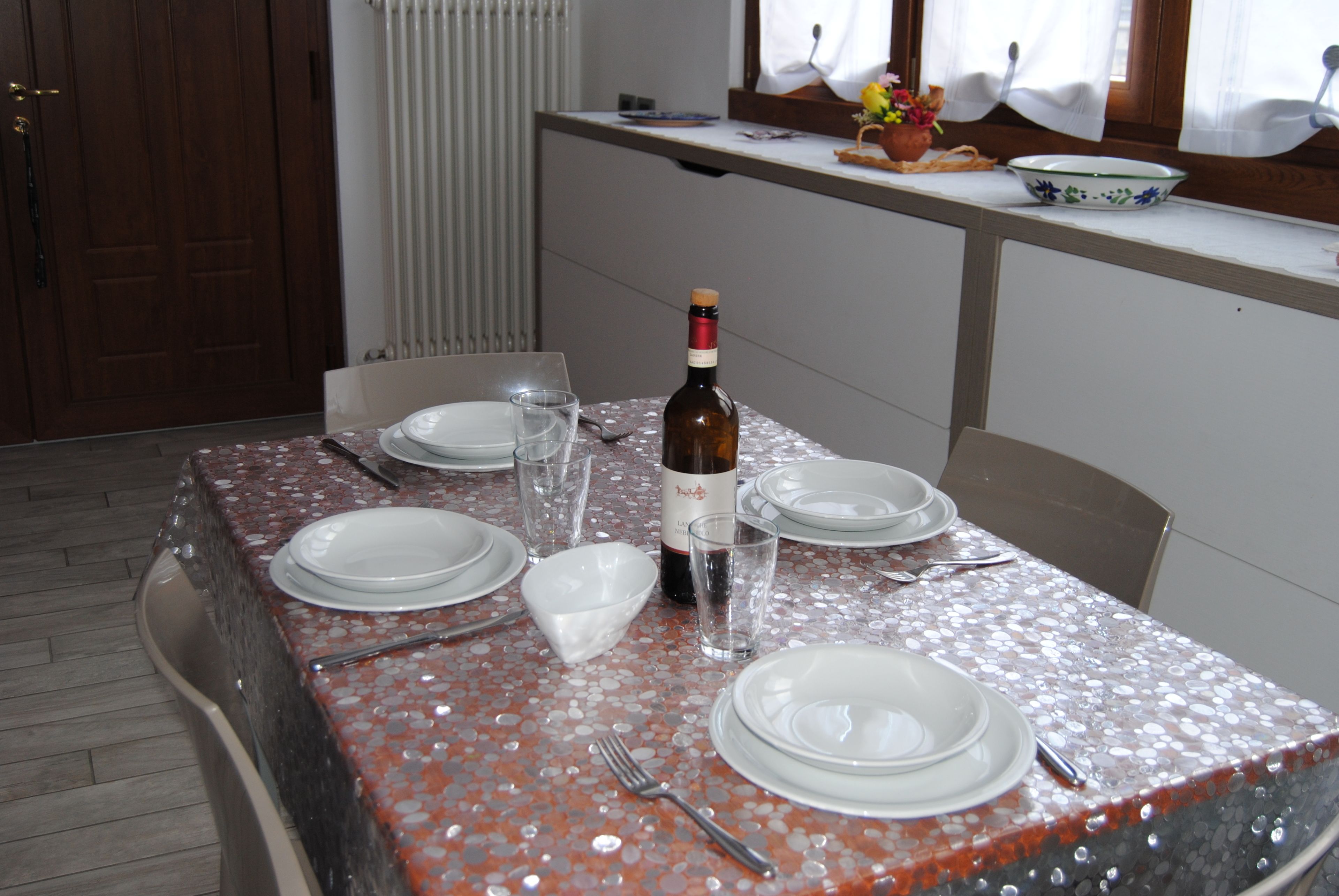Ein Esstisch ist gedeckt mit Tellern, Besteck und einer Weinflasche, über dem Tisch ein Fenster und dahinter eine Heizung.