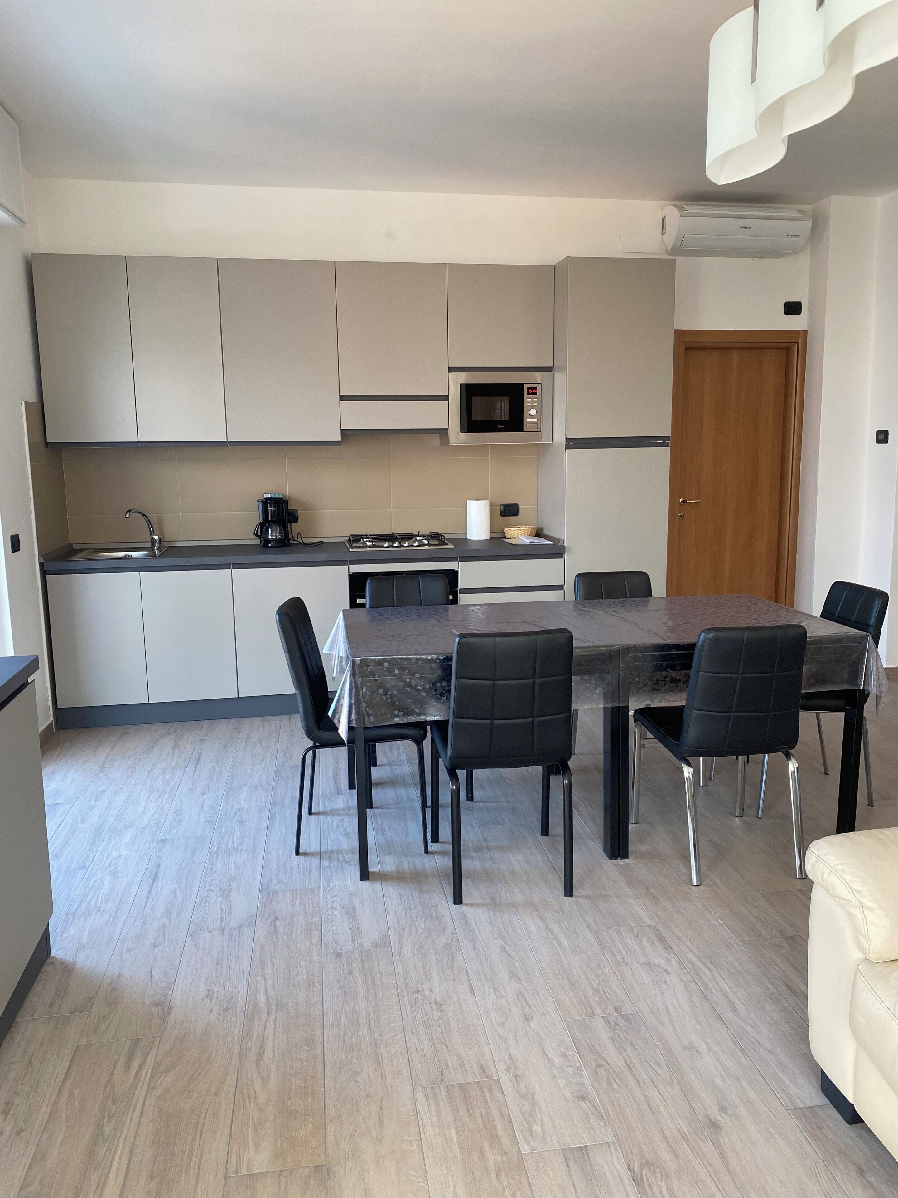 A modern kitchen with grey cabinets, a built-in microwave, and a stove. There is a dining table with black chairs and a beige sofa in the living area.