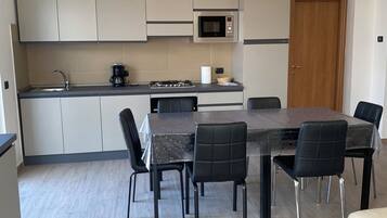 A modern kitchen with grey cabinets, a built-in microwave, and a stove. There is a dining table with black chairs and a beige sofa in the living area.