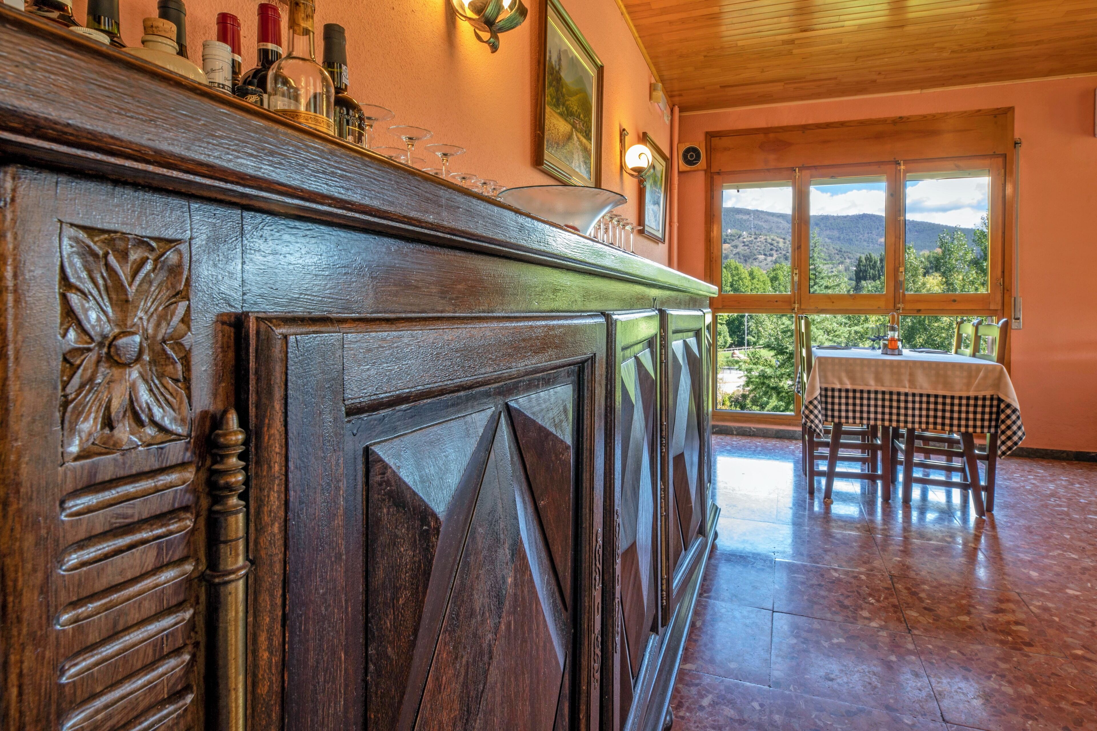 Een bar met een houten toonbank, wijnglazen en flessen. Achterin staat een tafel met stoelen, en er is uitzicht op bergen door een raam.