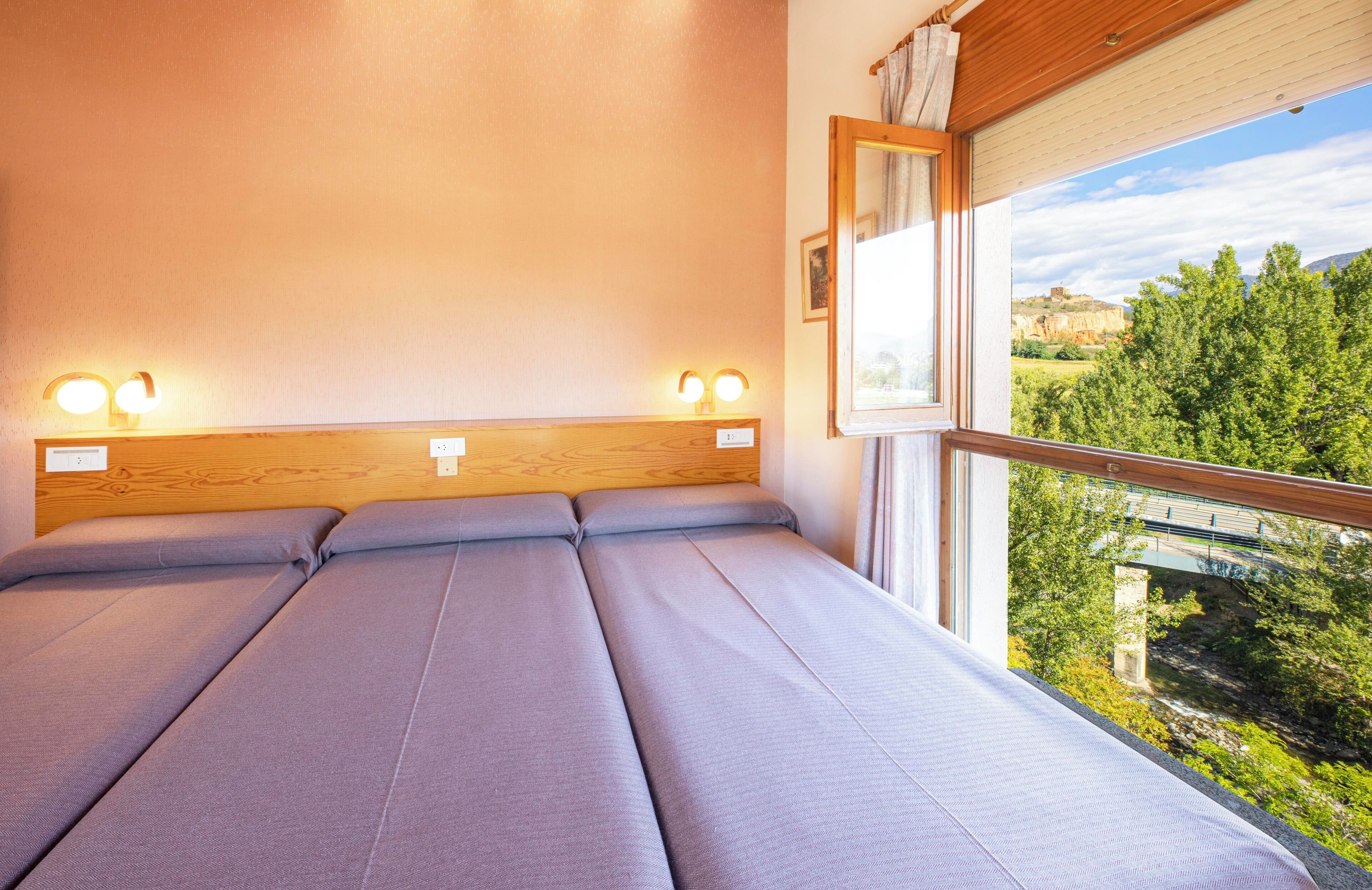 A room with a large bed, two bedside lamps, and a window offering a view of greenery and a distant building.