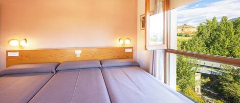 A room with a large bed, two bedside lamps, and a window offering a view of greenery and a distant building.