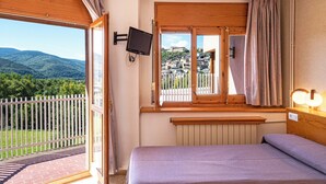 A bedroom with a bed, a TV mounted on the wall, a window with curtains, and a view of a mountainous landscape.