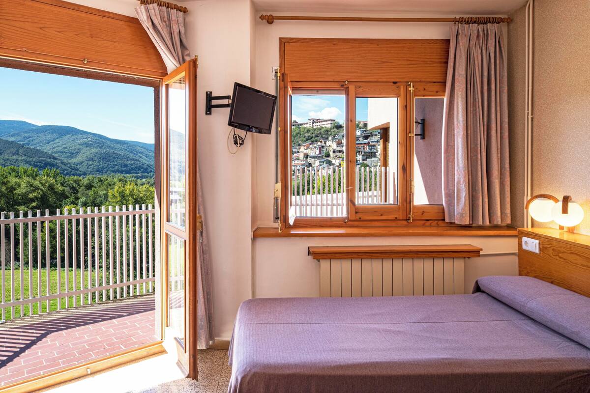 Un dormitorio con una cama, un televisor montado en la pared, una ventana con cortinas y vistas a un paisaje montañoso.