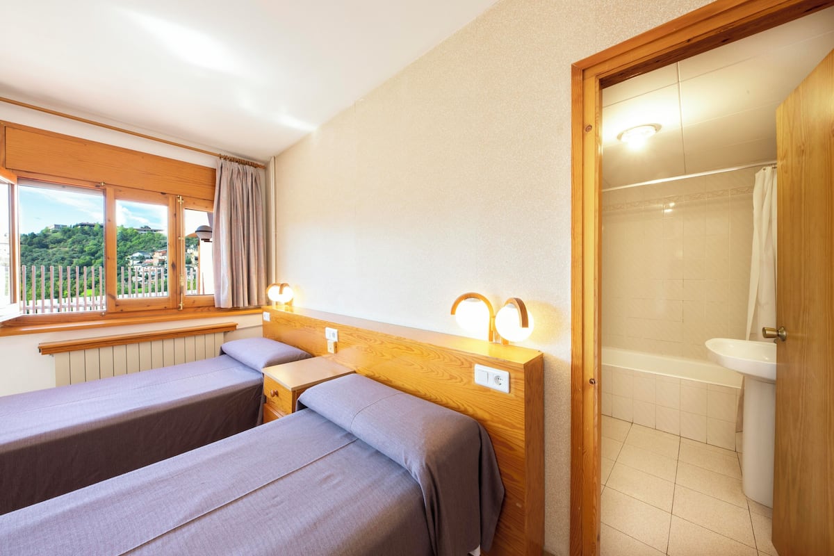Habitación con dos camas, una ventana con cortinas, cabecera de madera, baño con bañera y lavamanos, y vista a zonas verdes.