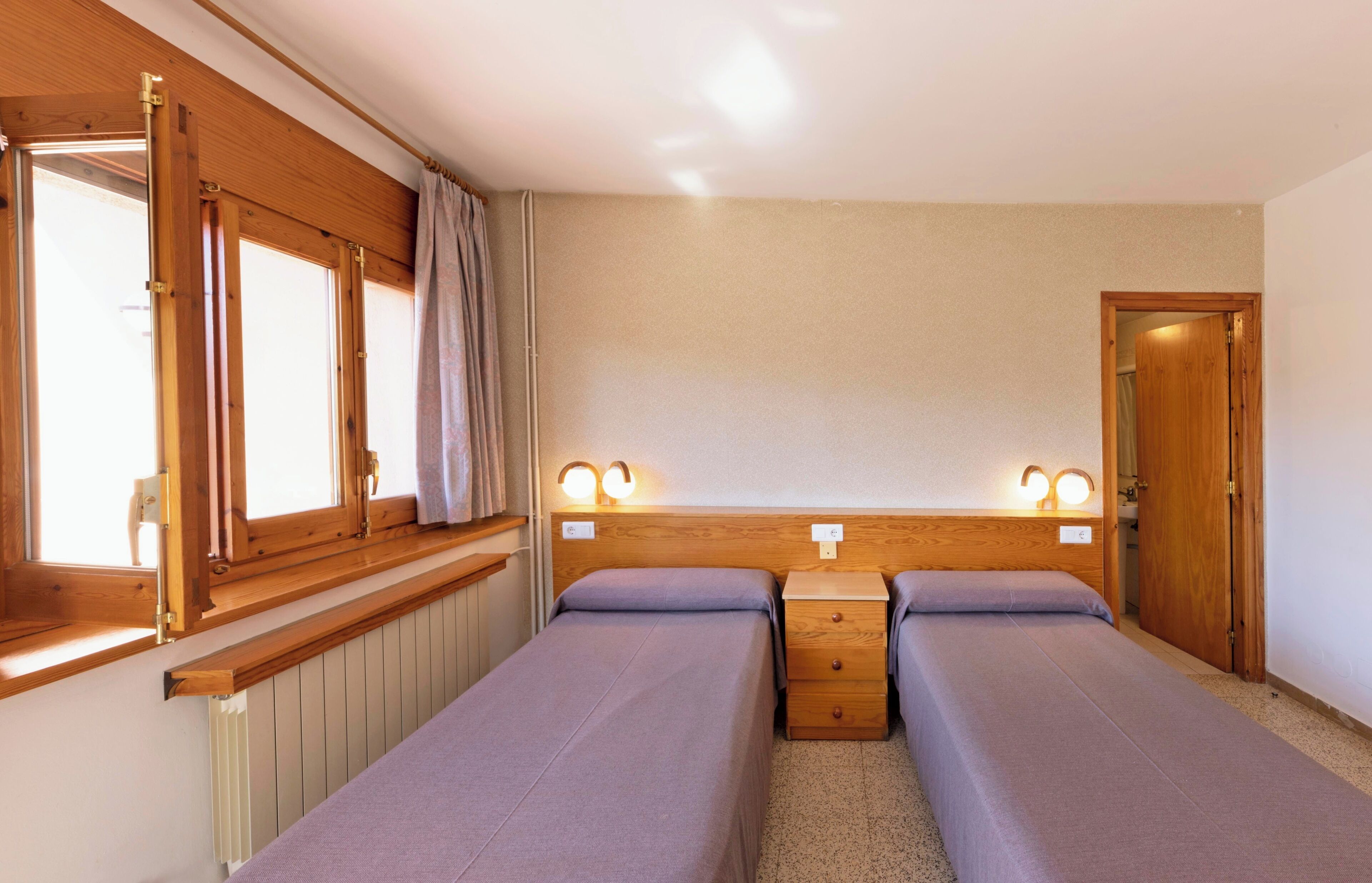 Une petite chambre propre, avec deux lits simples, des têtes de lit en bois, des lampes de chevet et une fenêtre avec des rideaux.