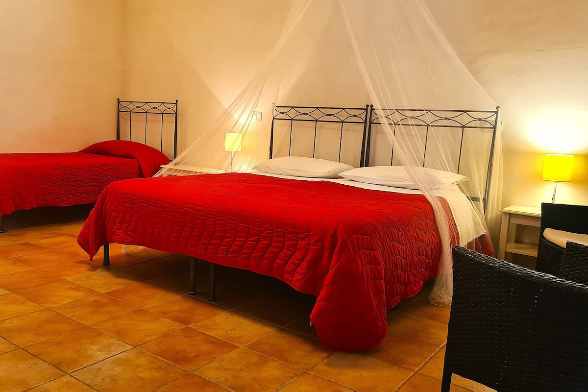 Ein Zimmer mit zwei Betten, jeweils mit roter Bettdecke und weißen Kissen, einem Moskitonetz über den Betten sowie einem kleinen Tisch mit Lampe.