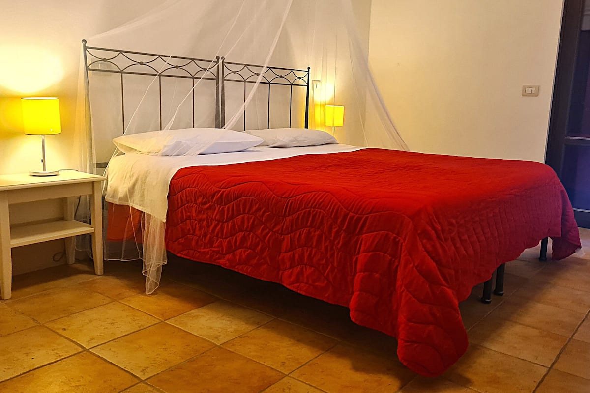 Ein Bett mit roter Bettdecke, zwei Nachttische mit Lampen und ein weißer, hockerförmiger Bettrahmen.