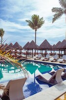 Zona junto a la piscina con sillas de descanso y sombrillas de paja, una palmera y cielo despejado.