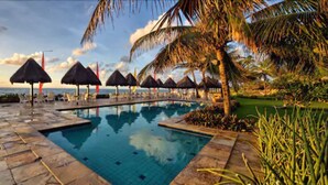 A pool area with lounge chairs and umbrellas, surrounded by palm trees and a view of the ocean.