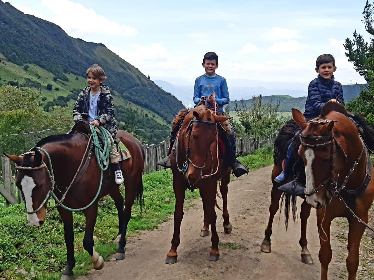 Three people riding horses on a dirt path with a mountainous backdrop.