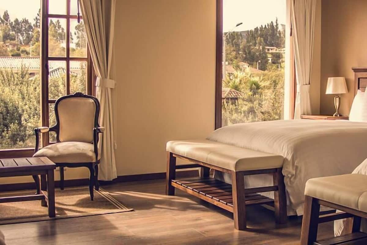 A bedroom with a large bed, a chair, and a bench. There is a window with a view of trees and buildings.