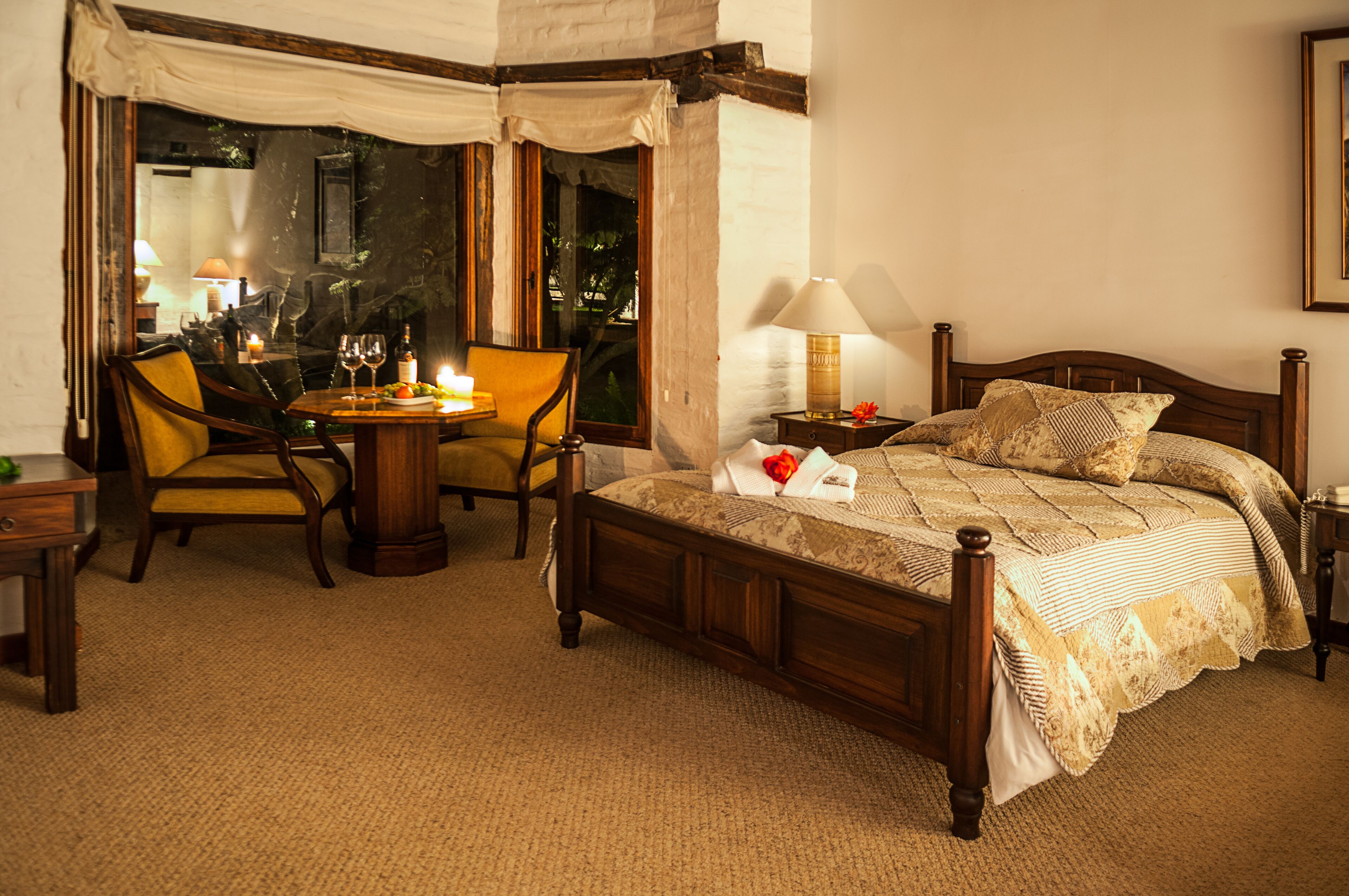 A hotel room with a wooden bed, a round dining table with chairs, a lamp, and a view through a large window.