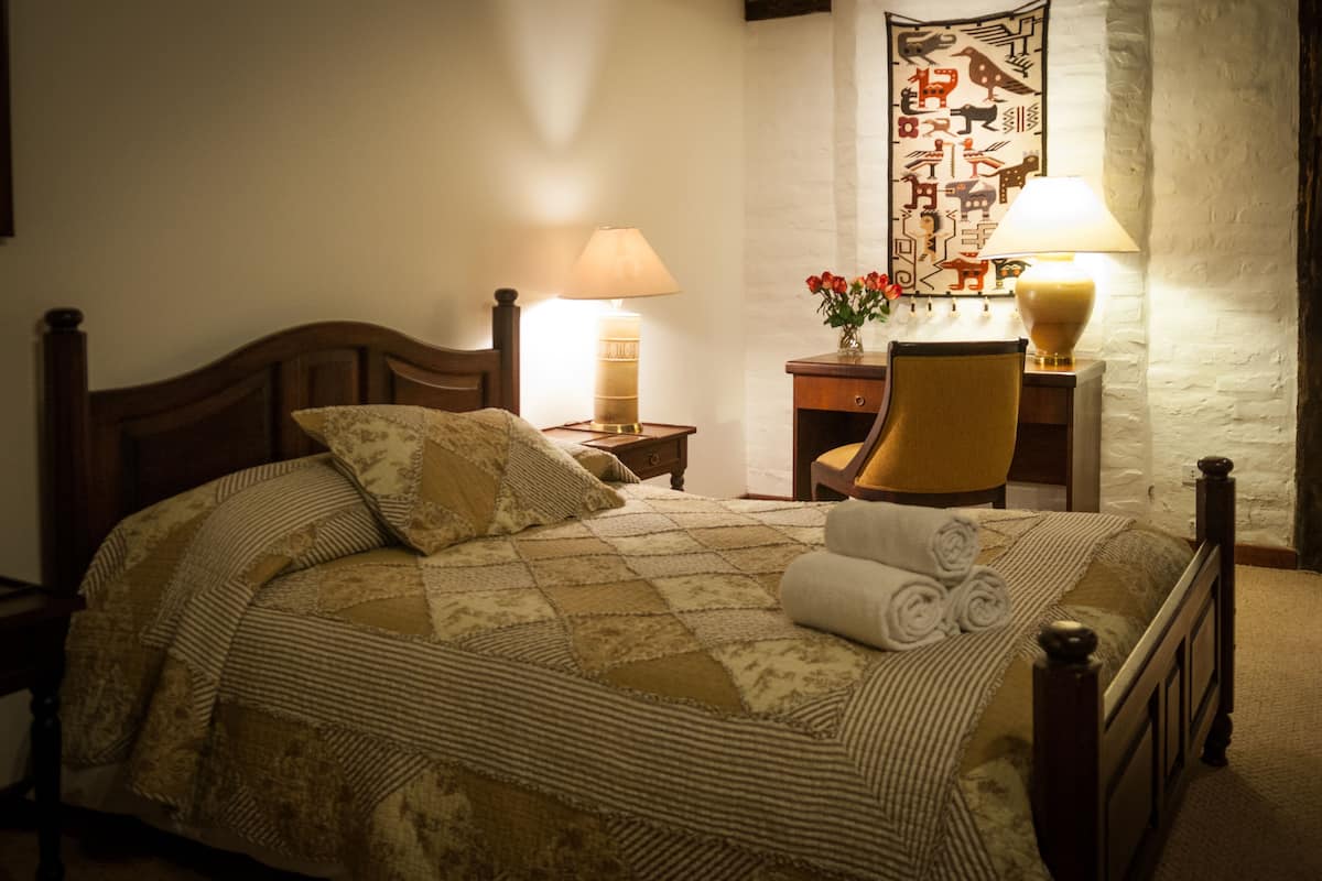 A neatly made bed with a quilt, two bedside lamps, a chair, and a wall tapestry.