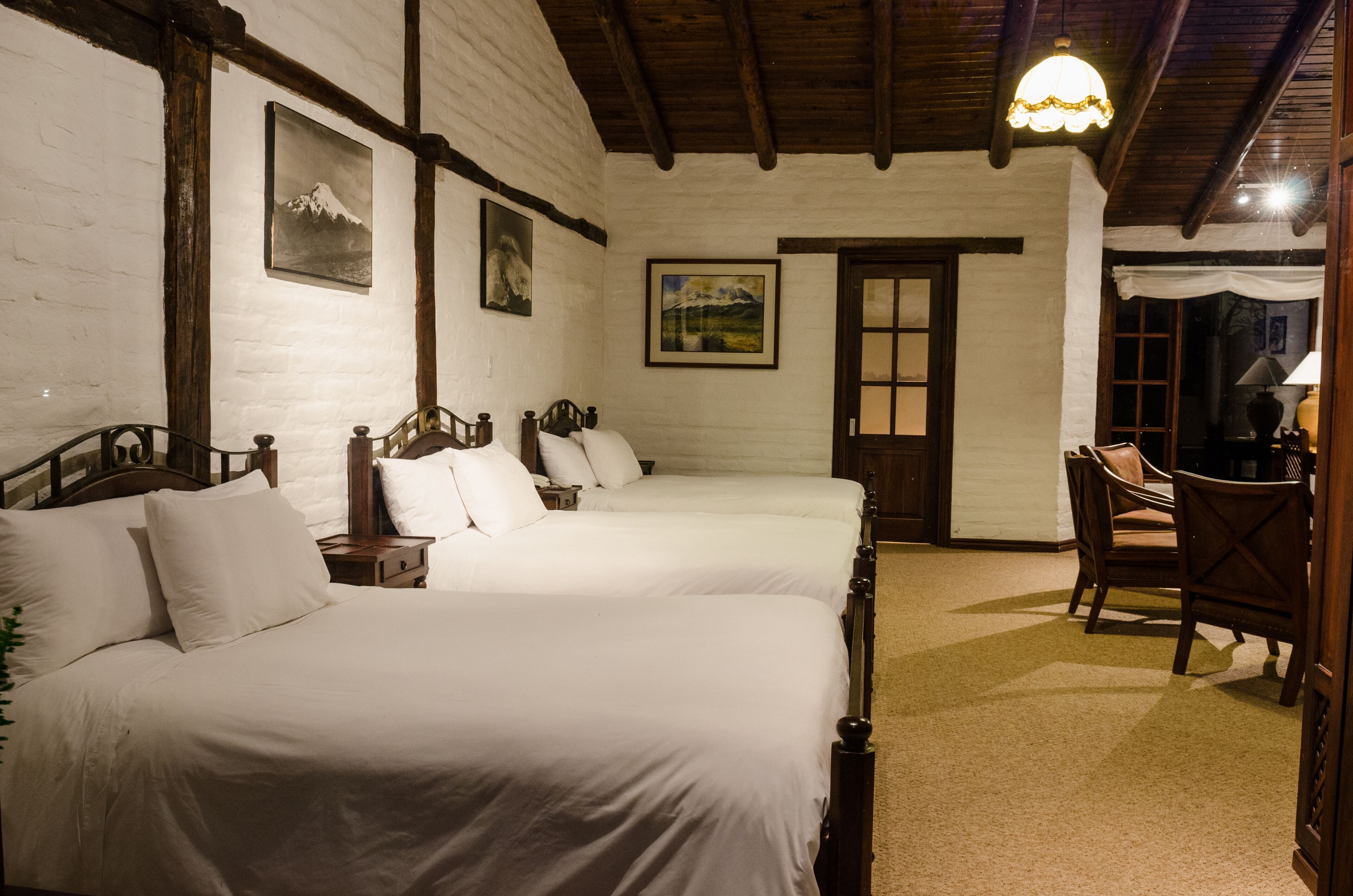 Una habitación con dos camas, cabeceros de madera, ropa de cama blanca, una silla de madera y una puerta con ventanal.