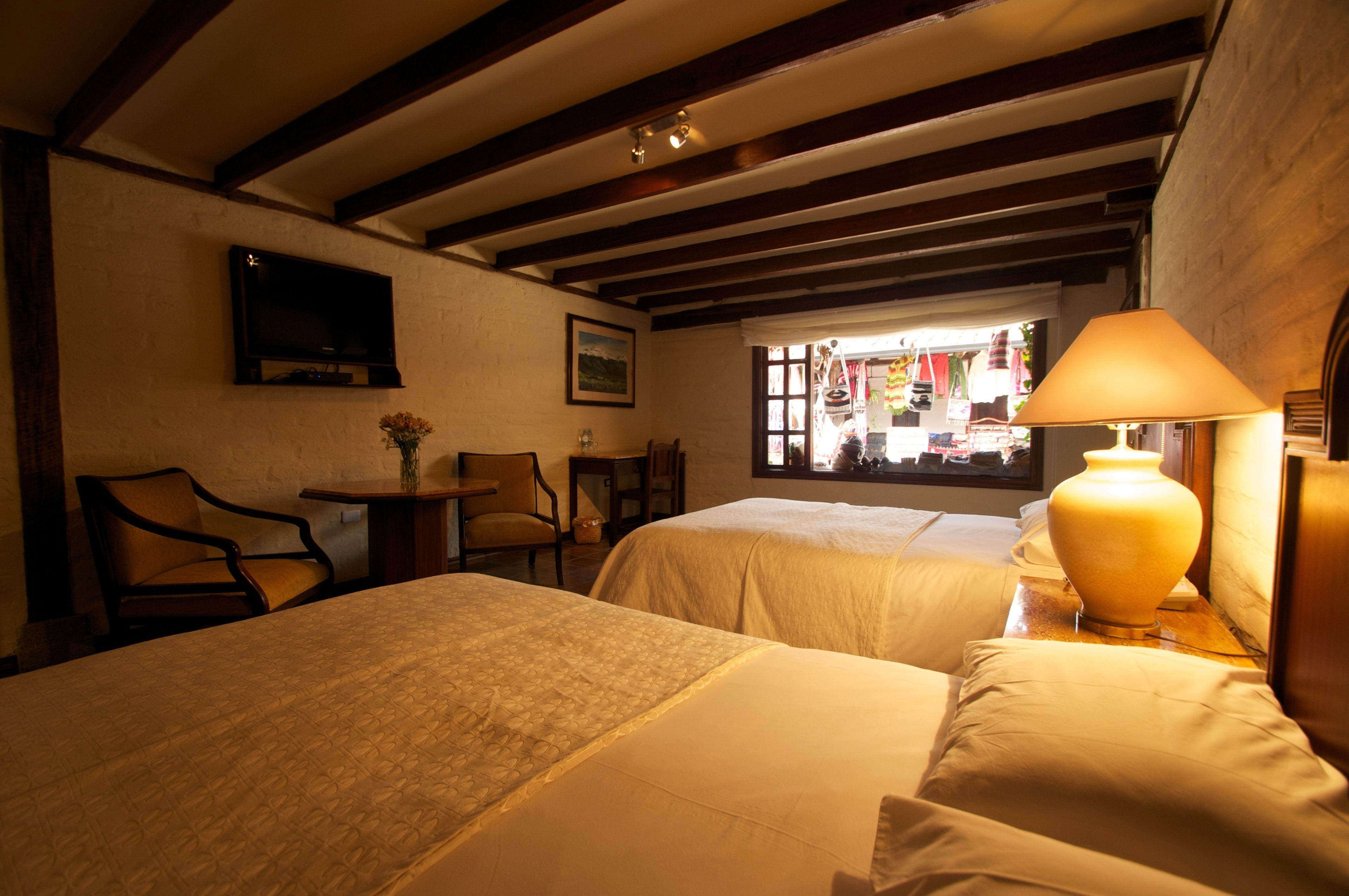 A hotel room with two beds, a TV mounted on the wall, a window with a view of the street, and a lamp on the nightstand.