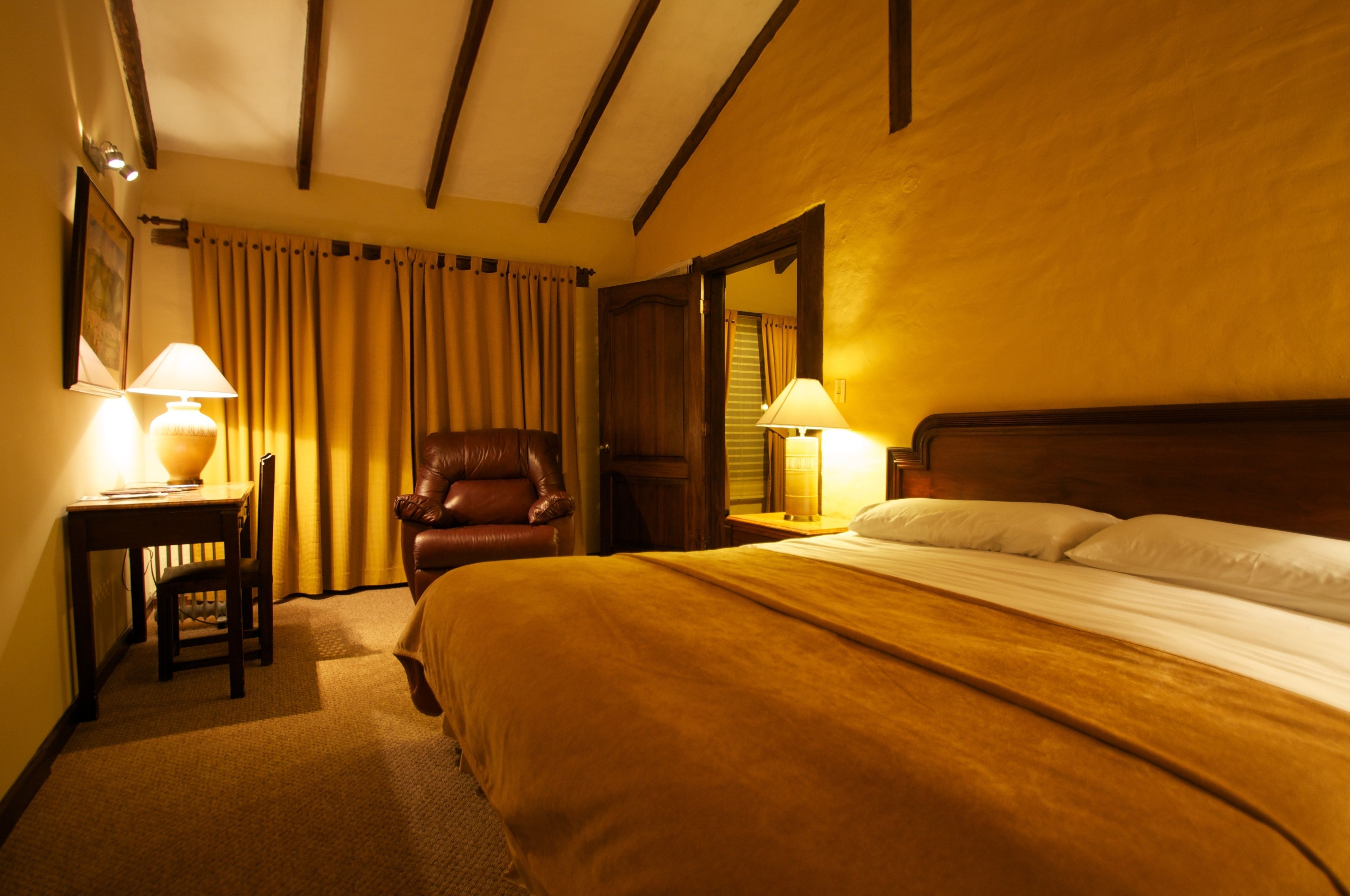 A hotel room with a large bed, two bedside lamps, a wooden headboard, a leather armchair, and a wooden desk with a lamp.