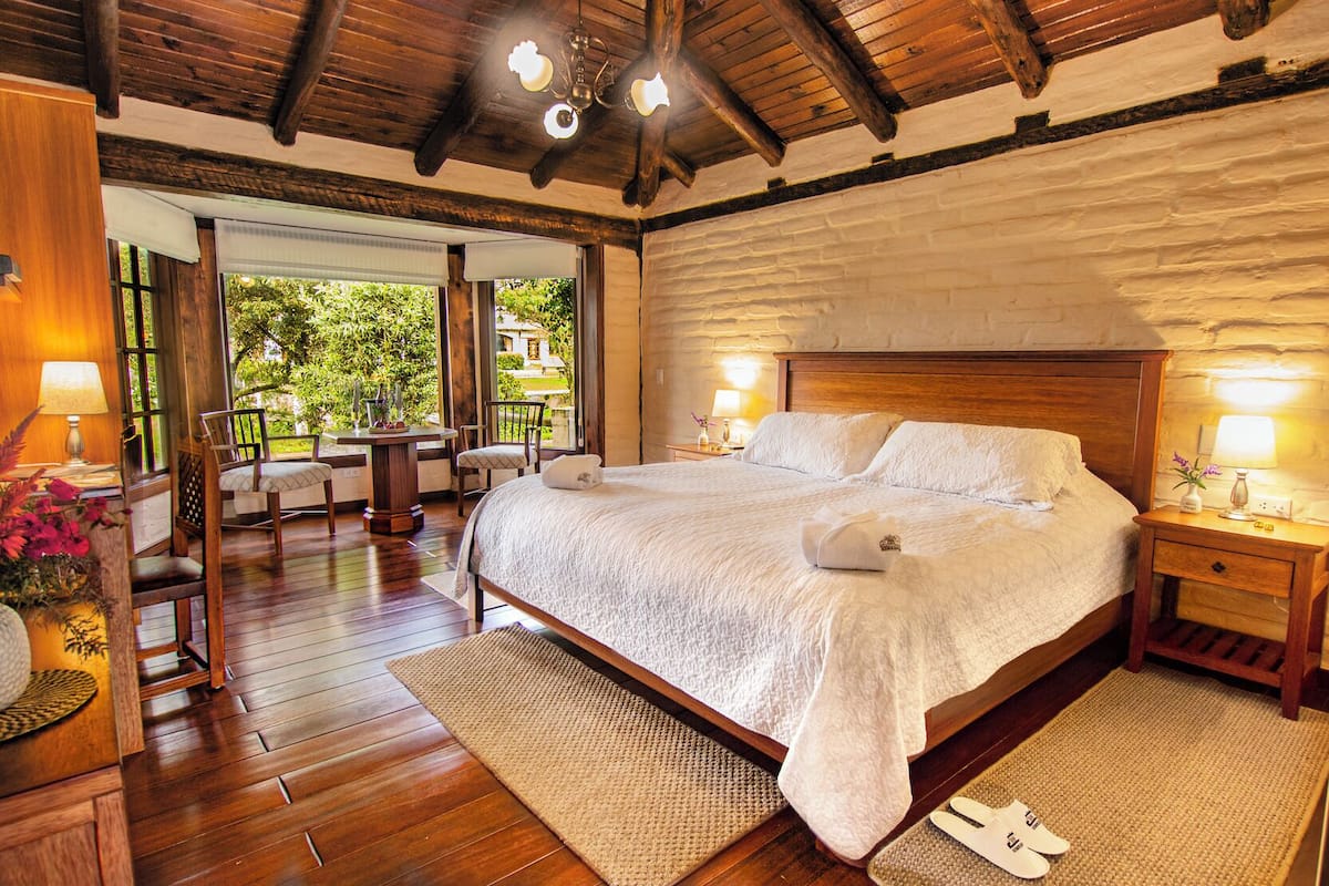 A bedroom with a large bed, wooden flooring, a stone wall, and a view of greenery outside.